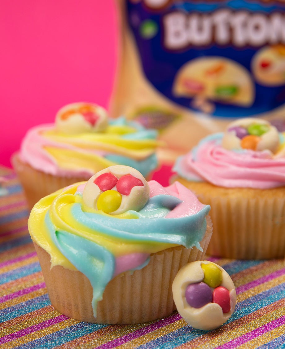 Close up of vanilla cupcakes with pink, blue and white swirls of icing topped with colourful Smarties buttons