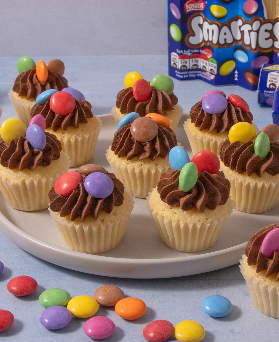 A tray of mini vanilla cupcakes with chocolate buttercream swirls and dotted with Smarties for decoration
