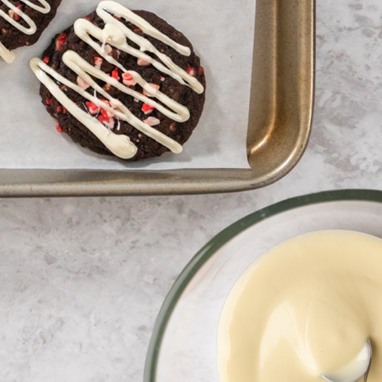 Glass bowl of white chocolate ganache with a metal dessertspoon in it, pictured next to three dark chocolate cookies