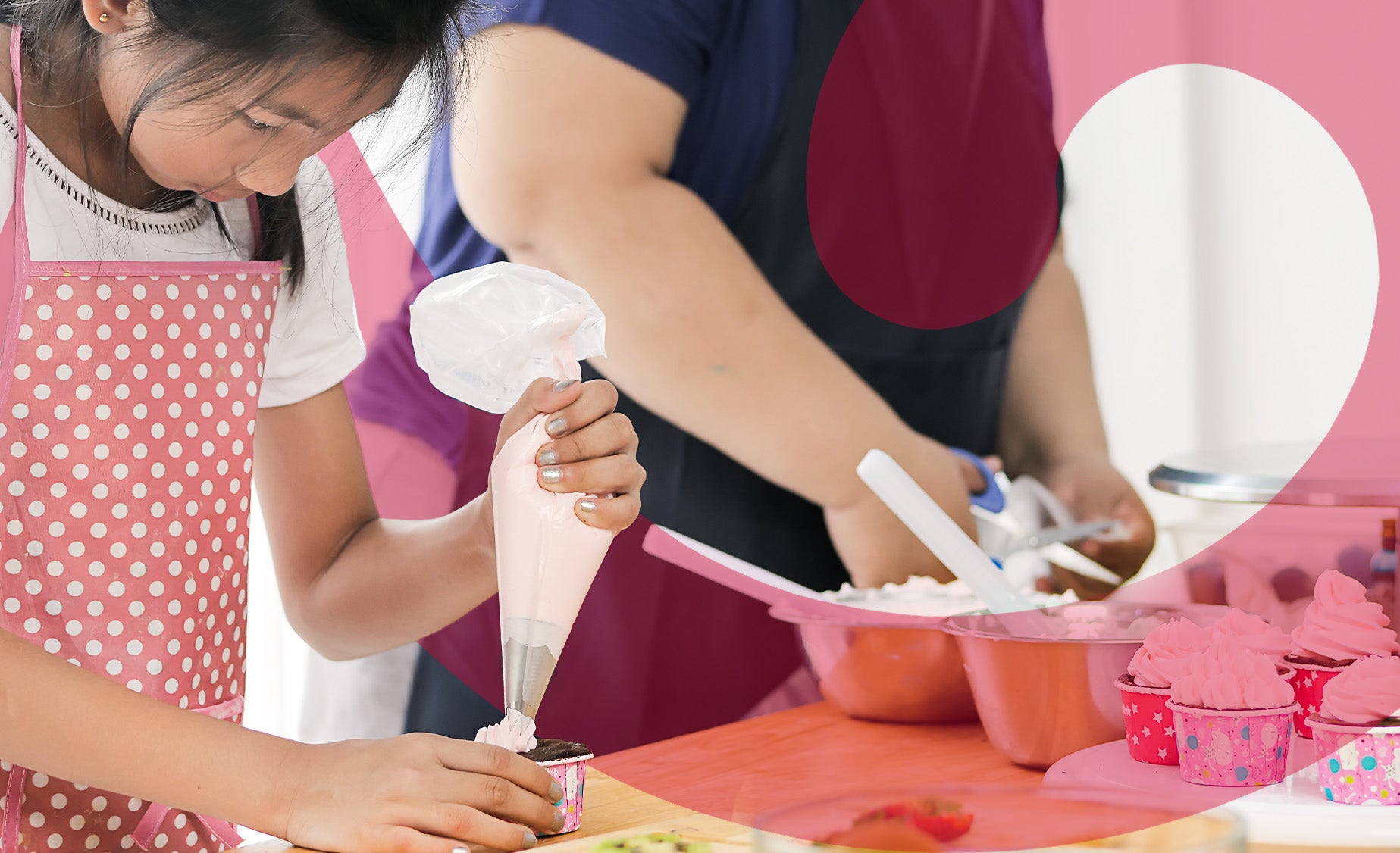 A girl in a pink and white spotted apron decorates a cupcake on a table with an adult beside her.