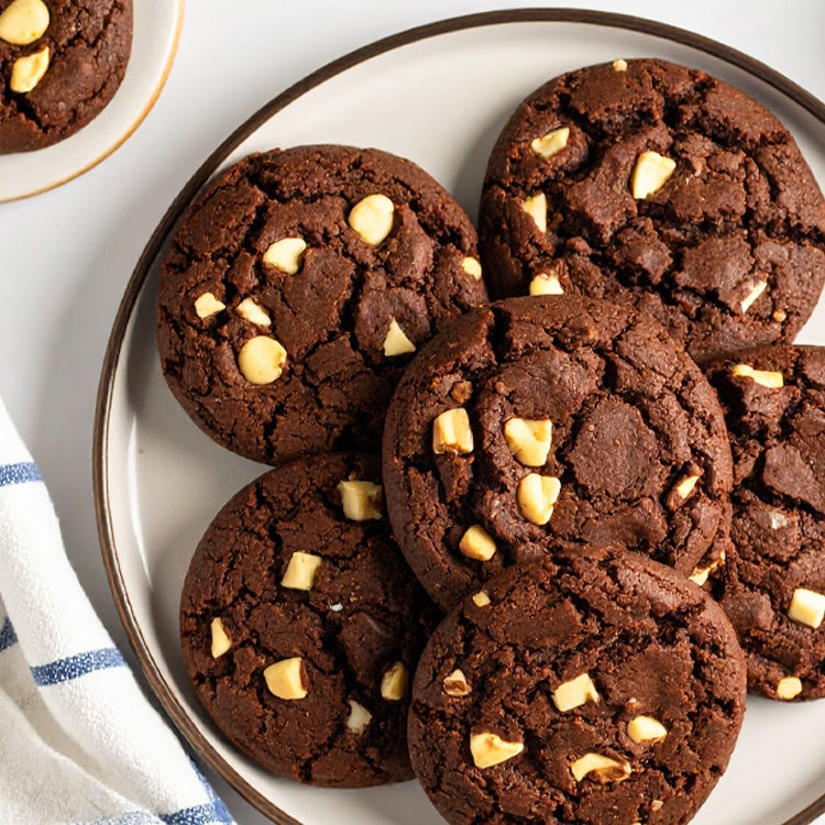 Chocolate cookies with white Milkybar chocolate chunks on a plate, surrounded by a linen kitchen towel and extra white chocol