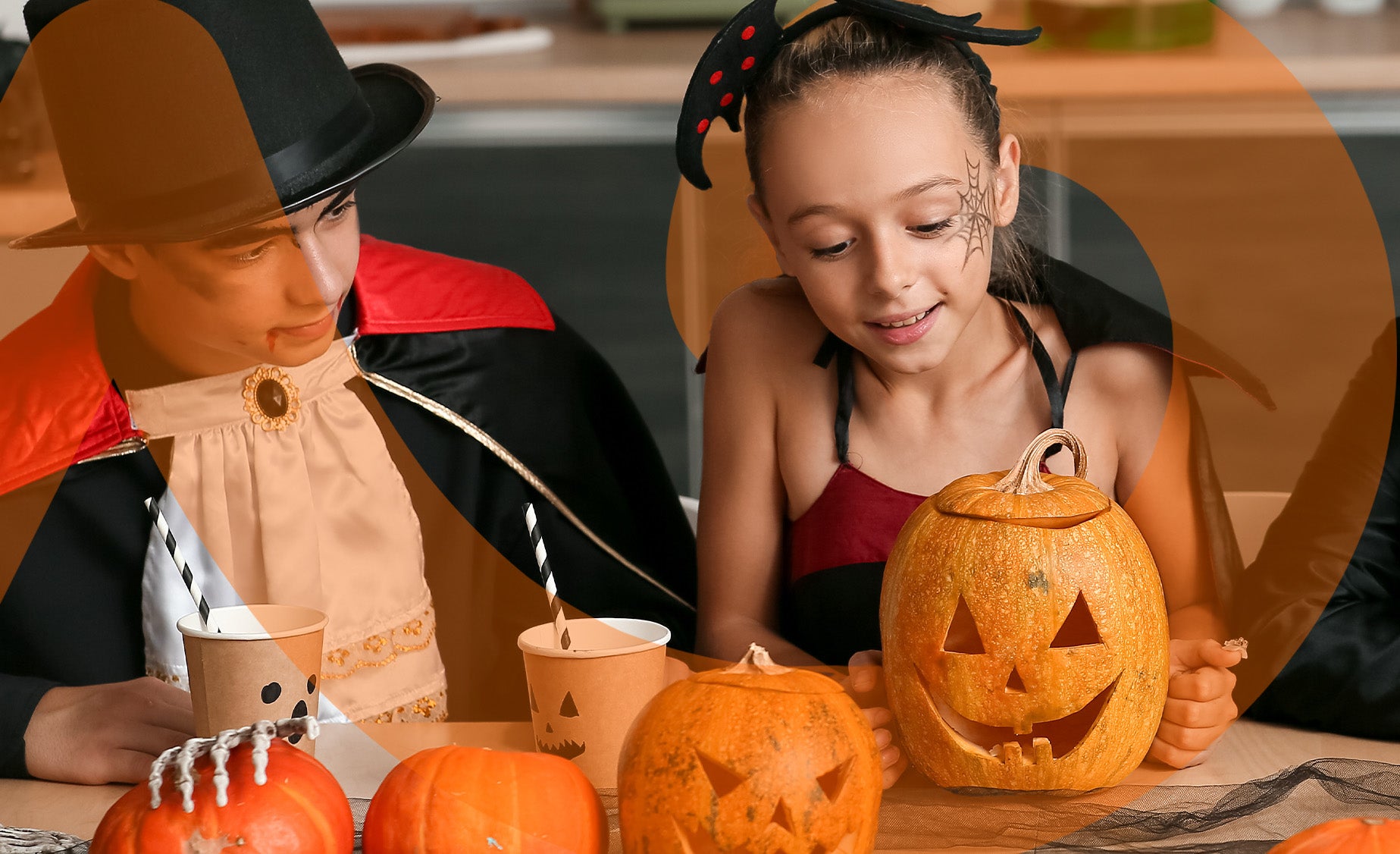 A boy and a girl dressed in Halloween costumes sat at a table with carved pumpkins and drinks with paper straws.