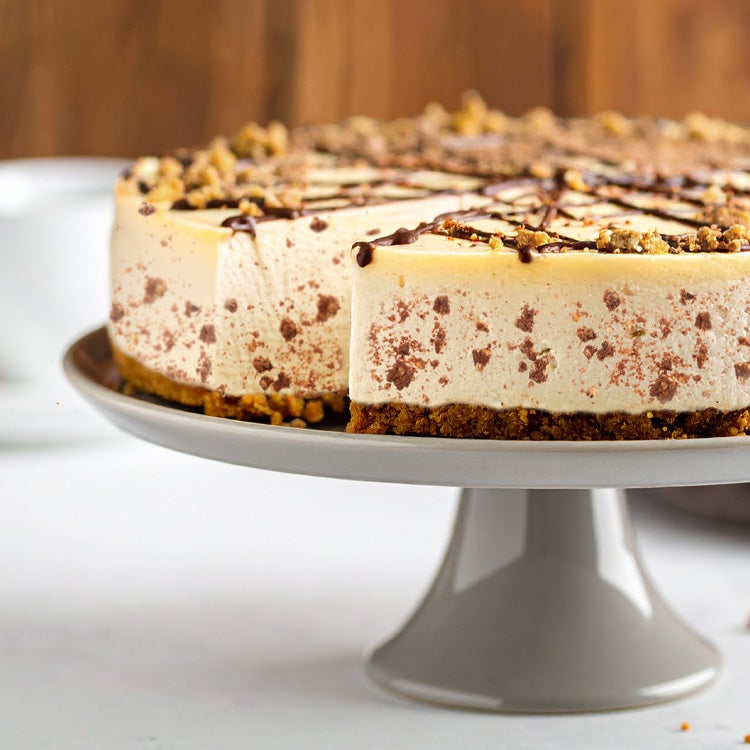 Toffee crisp cheesecake on a cake stand with a missing slice appearing on a dessert plate, on a grey table with a coffee cup 