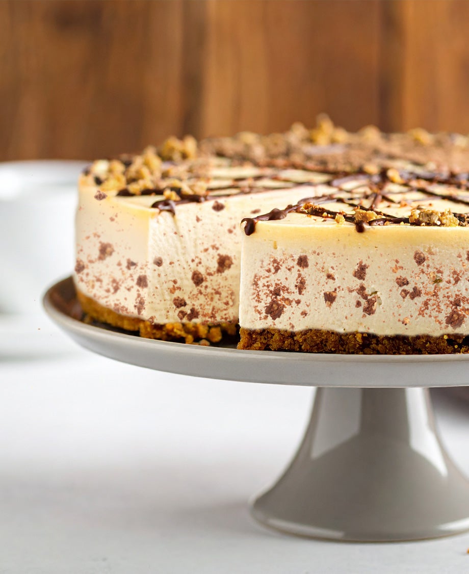 Toffee crisp cheesecake on a cake stand with a missing slice appearing on a dessert plate, on a grey table with a coffee cup