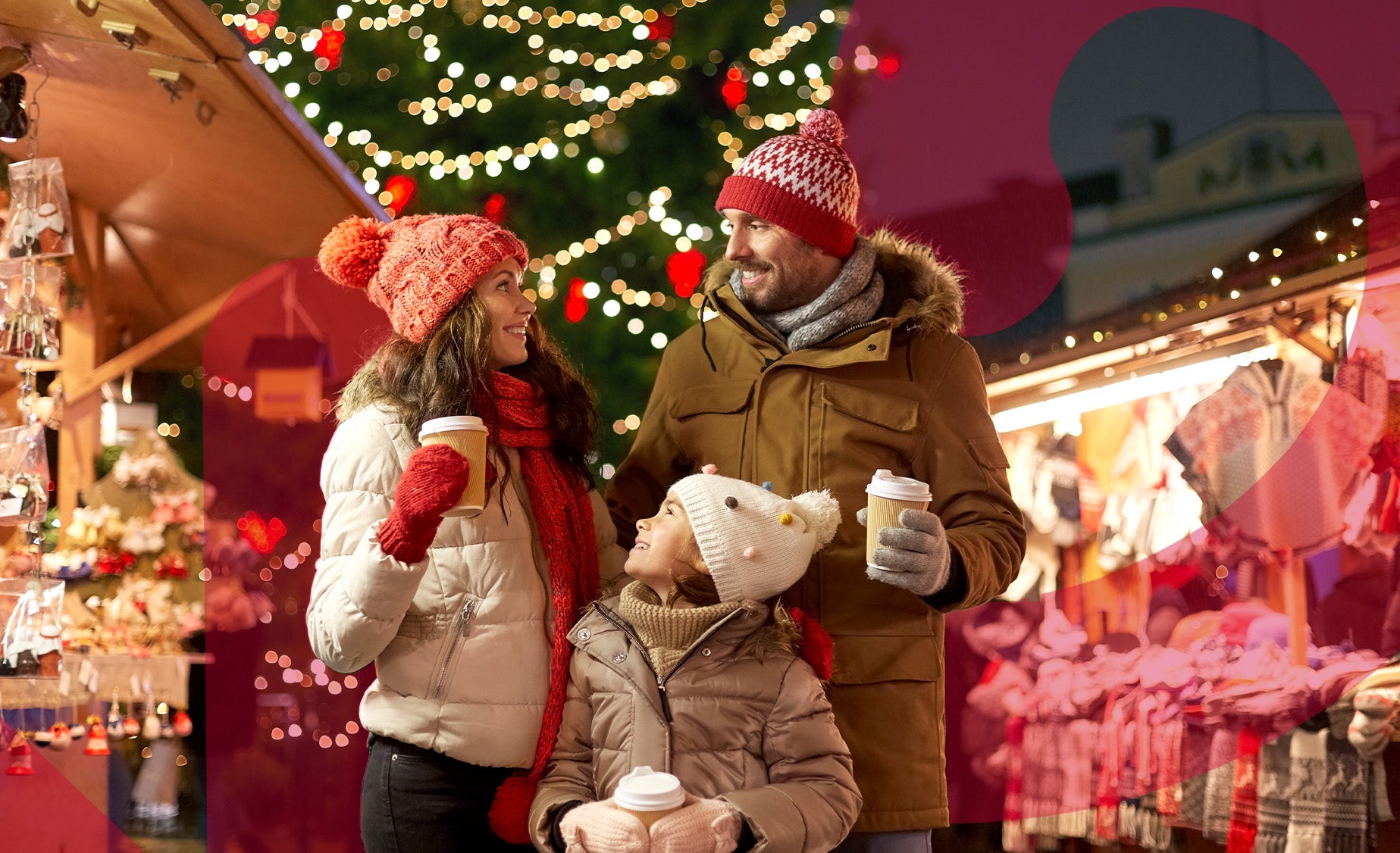 A couple and a child stand together in front of a festive Christmas market, surrounded by holiday decorations and lights.