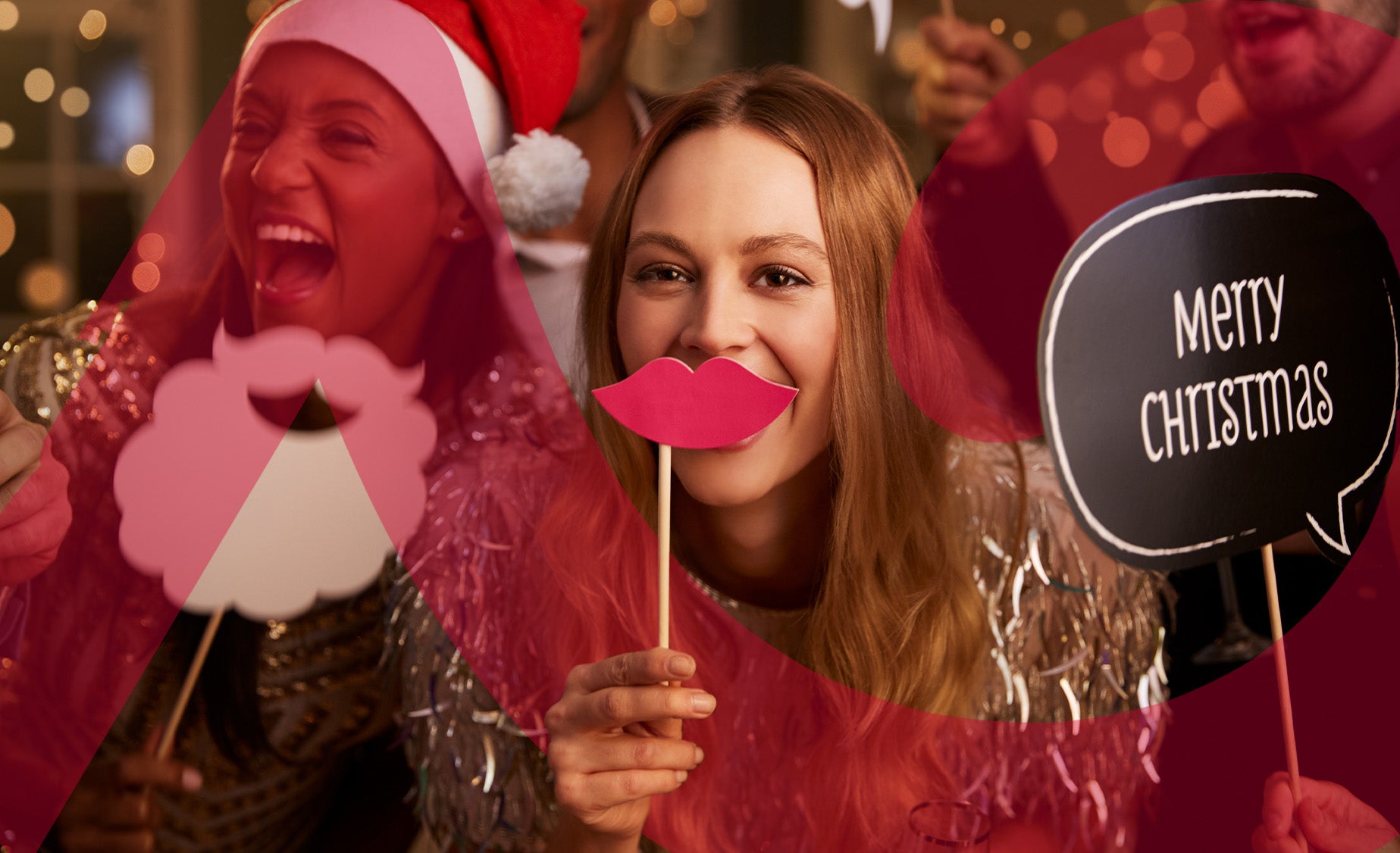 Two friends laughing together at Christmas with tinsel and a santa hat, with one holding a paper cut-out of joke lips to her 