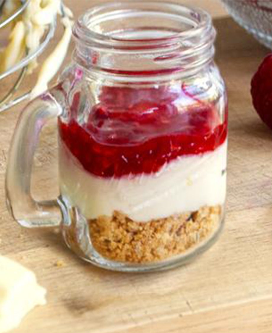 Glass jar with a chunky handle, containing layers of cheesecake base, creamy filling and raspberry coulis topping