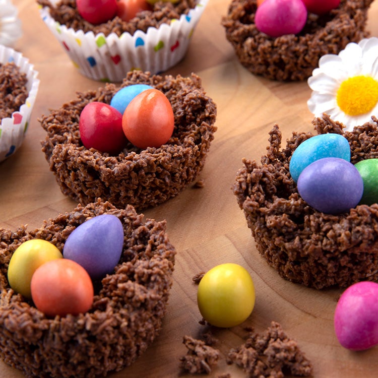  Chocolate cornflake nests, filled with colourful Smarties Mini Eggs, set on a wooden surface with a white daisy decoration.