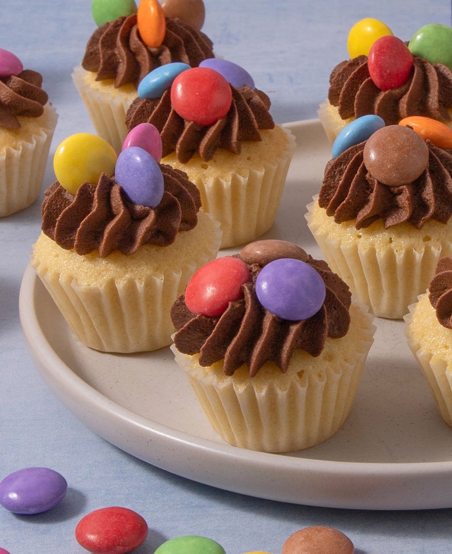 Mini vanilla cupcakes on a white plate, with chocolate buttercream piped on top and three Smarties on each one