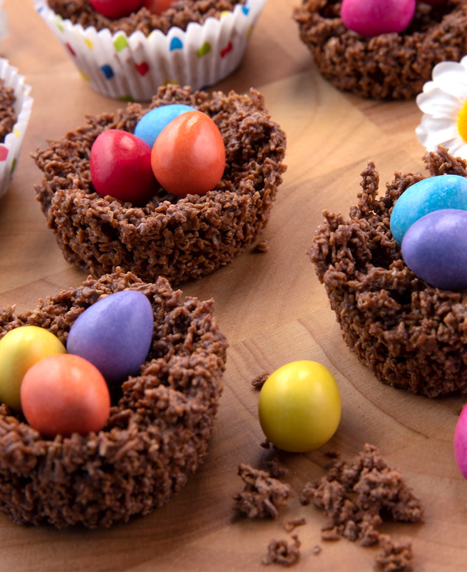Chocolate cornflake nests, filled with colourful Smarties Mini Eggs, set on a wooden surface with a white daisy decoration.