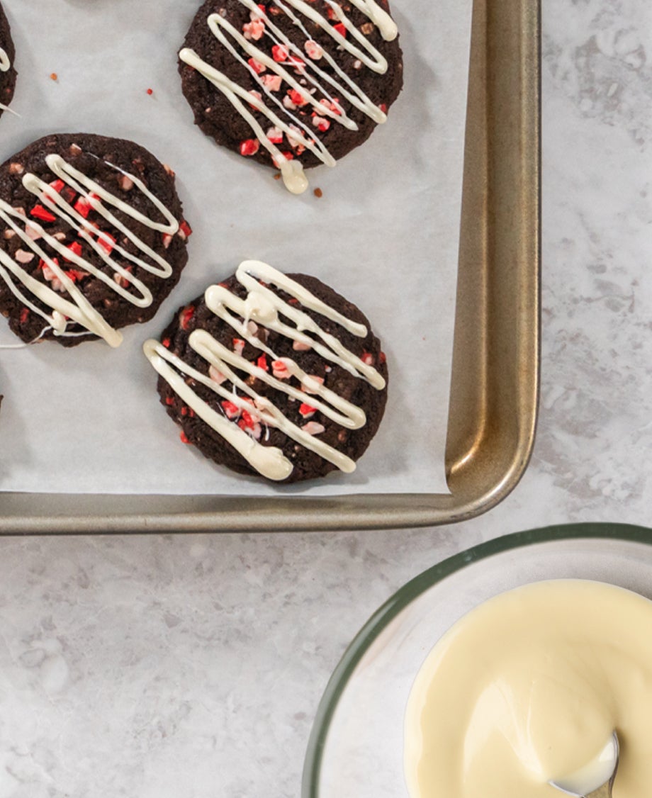Glass bowl of white chocolate ganache with a metal dessertspoon in it, pictured next to three dark chocolate cookies