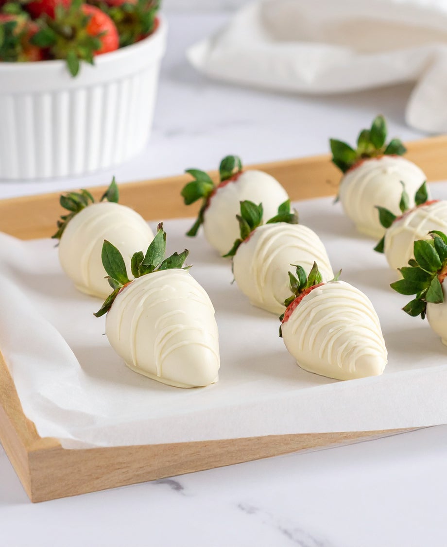 Fresh strawberries dipped in white chocolate on a wooden tray with more fresh strawberries scattered around    