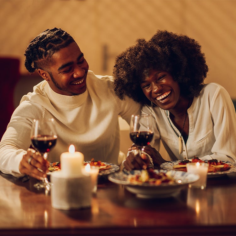 A couple laugh together at dinner at home, with candles and glasses of red wine 