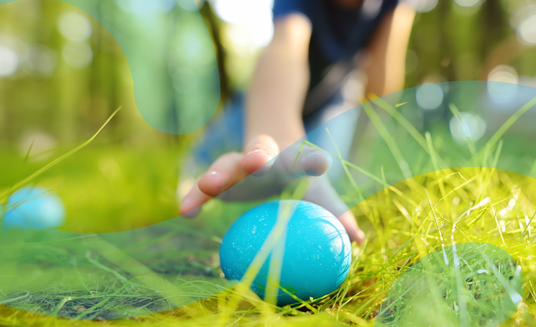 A child’s hand reaching for a colourful blue Easter egg in the grass. 
