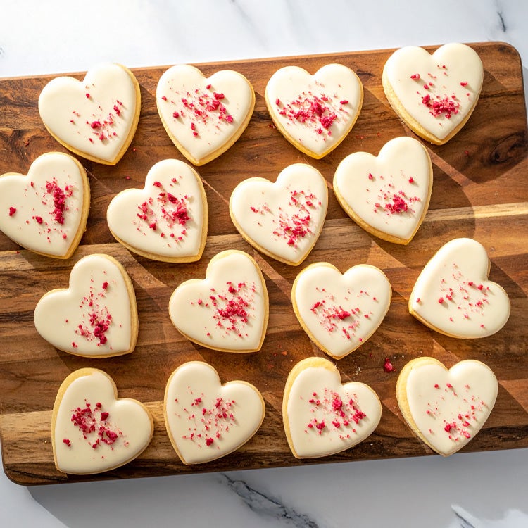 Batch of heart-shaped cookies topped with white chocolate icing scattered with dried raspberries, on a wooden board