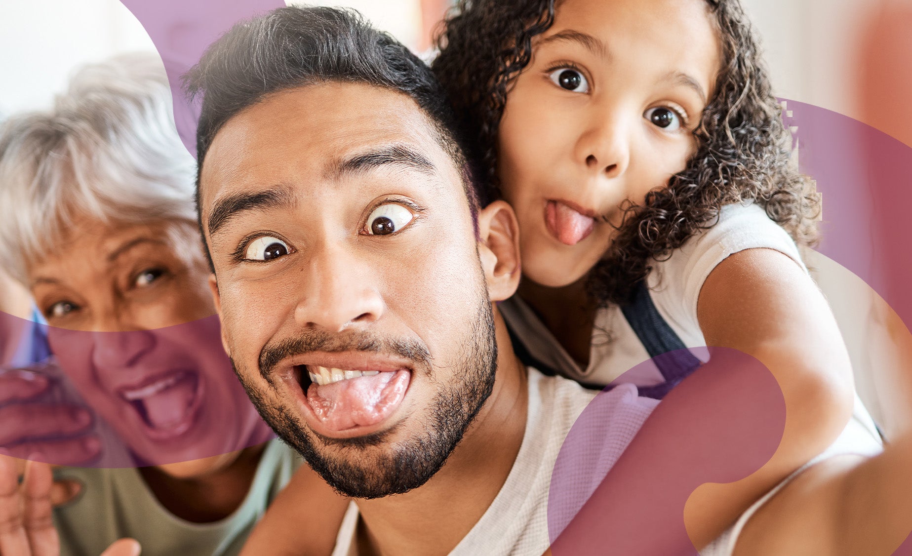 A father pulling a silly face while holding his daughter, who sticks out her tongue, with his mother in the background laughi