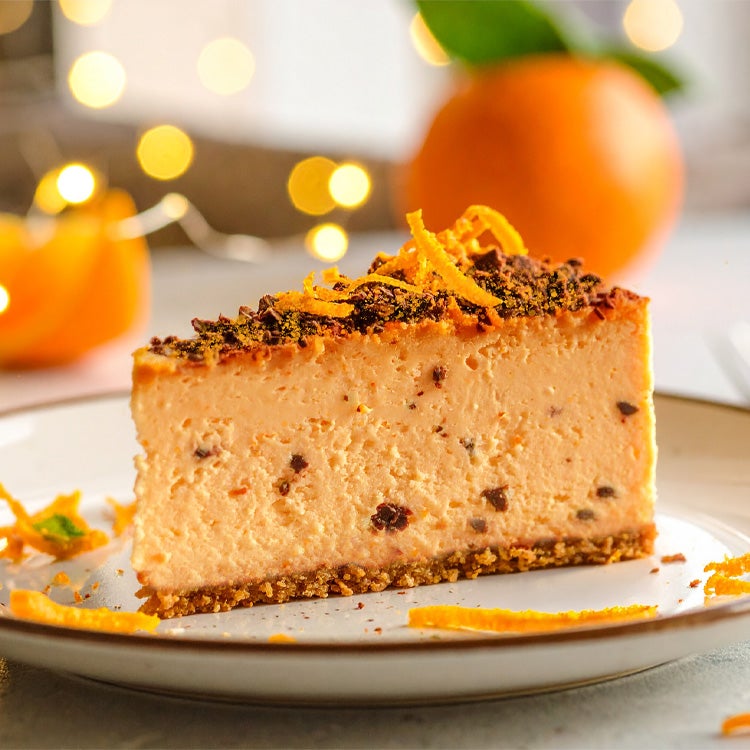 Slice of chocolate orange cheesecake, topped with crumbled Aero Orange and orange zest, on a white plate with oranges
