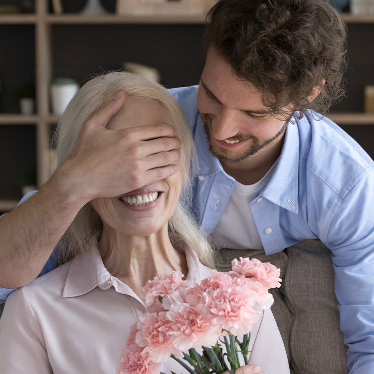 A smiling woman sits on a sofa with her eyes covered by a man, who is about to surprise her with a bunch of pretty carnations
