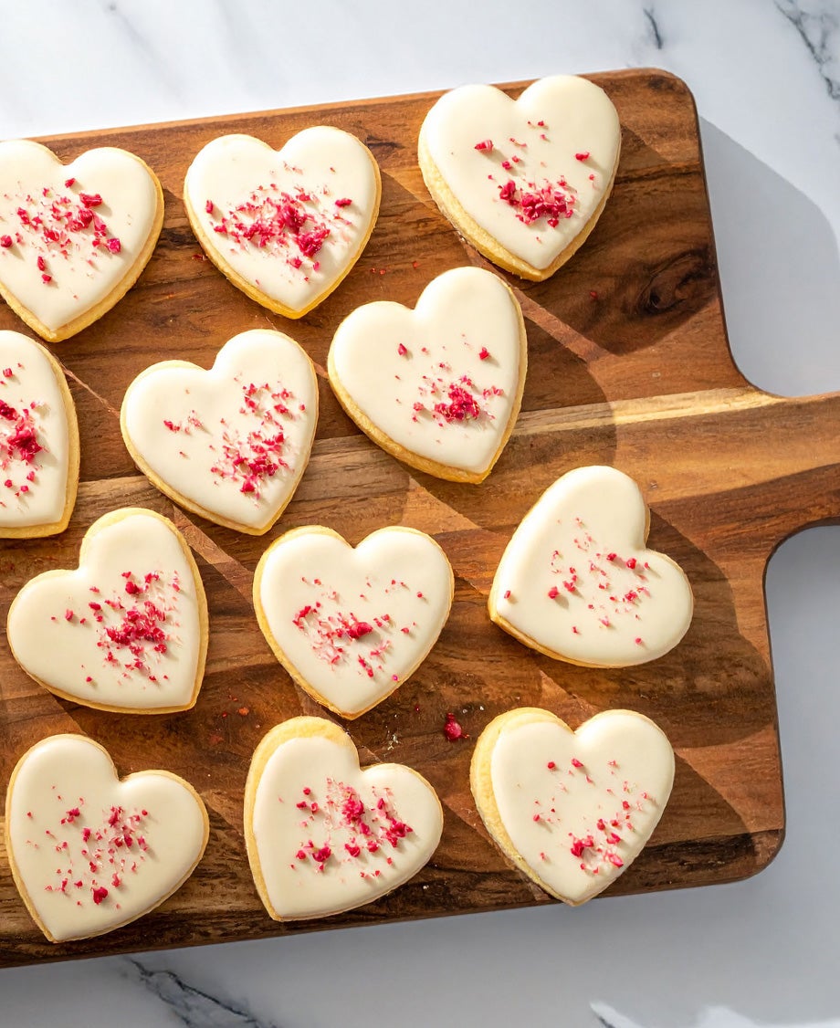 Batch of heart-shaped cookies topped with Milkybar white chocolate icing scattered with dried raspberries, on a wooden boar
