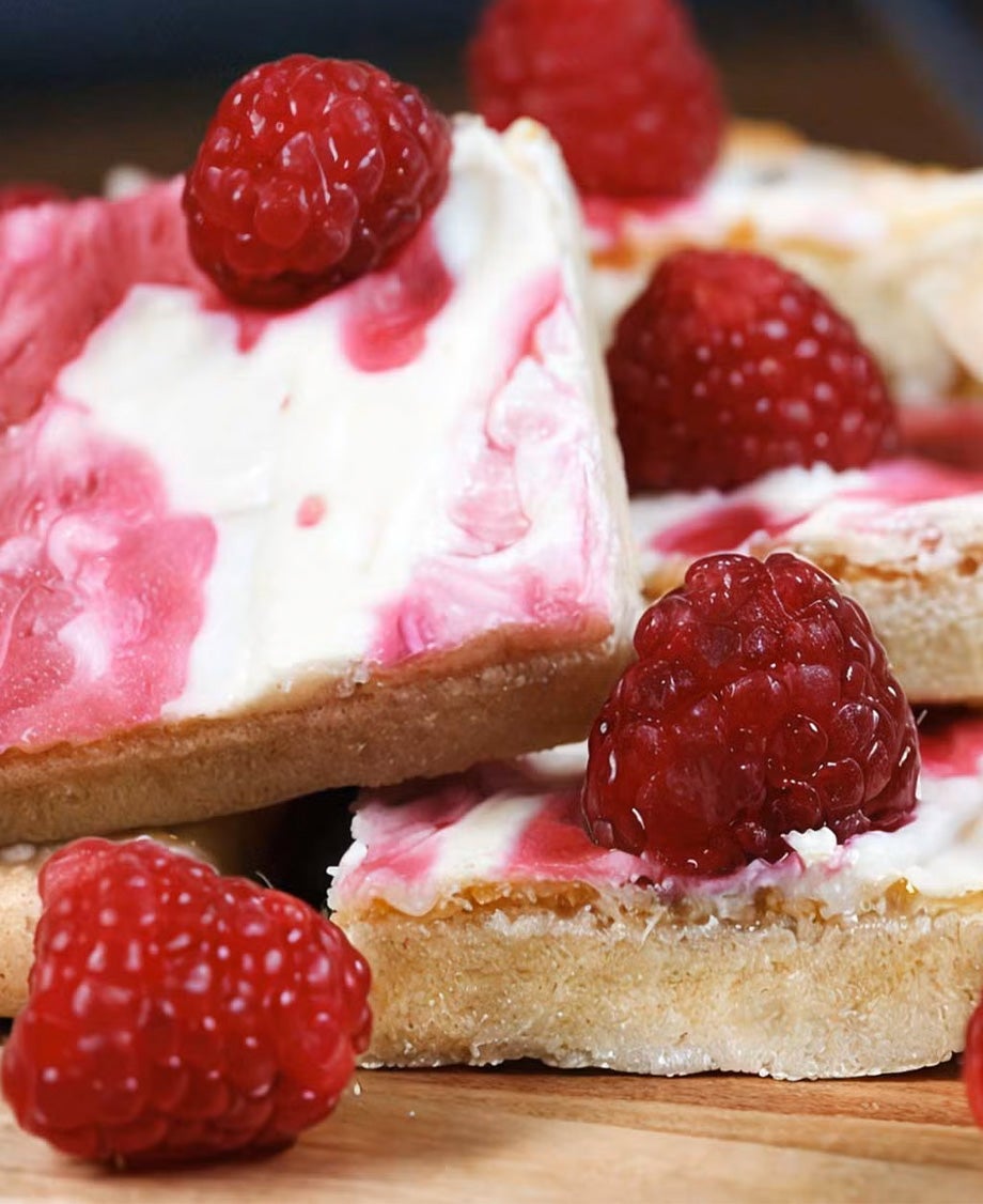  Squares of pastry topped with white chocolate and raspberry icing and fresh raspberries on a wooden worktop