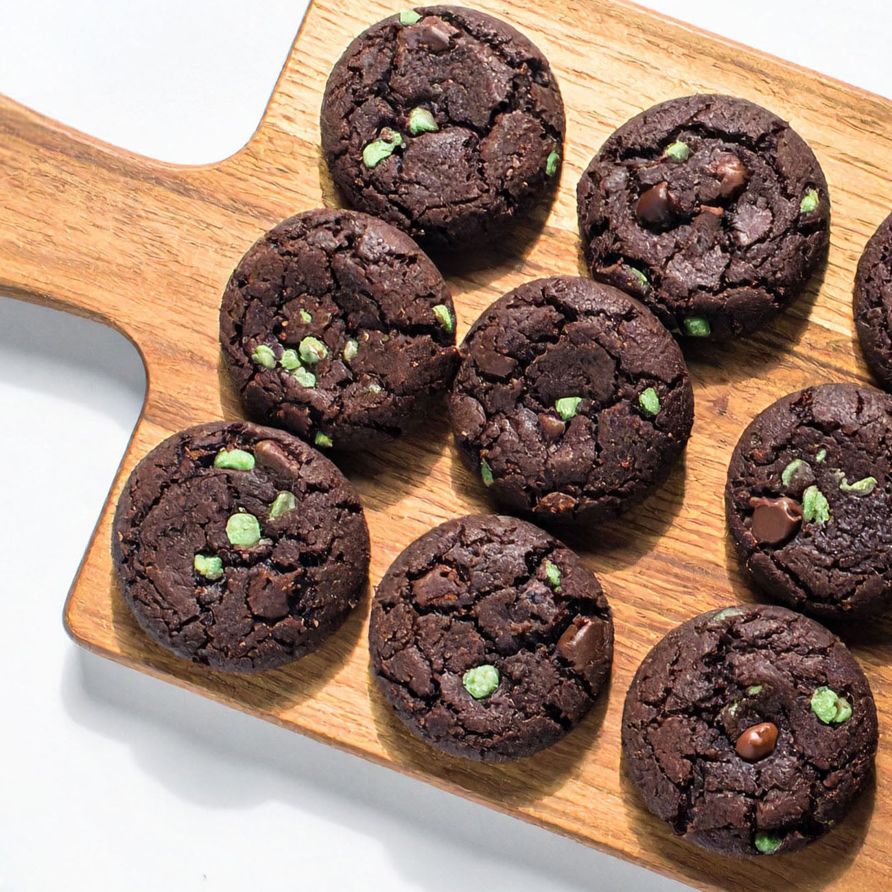 A wooden cutting board displays a stack of Mint chocolate chip cookies, showcasing their rich, dark color and texture.